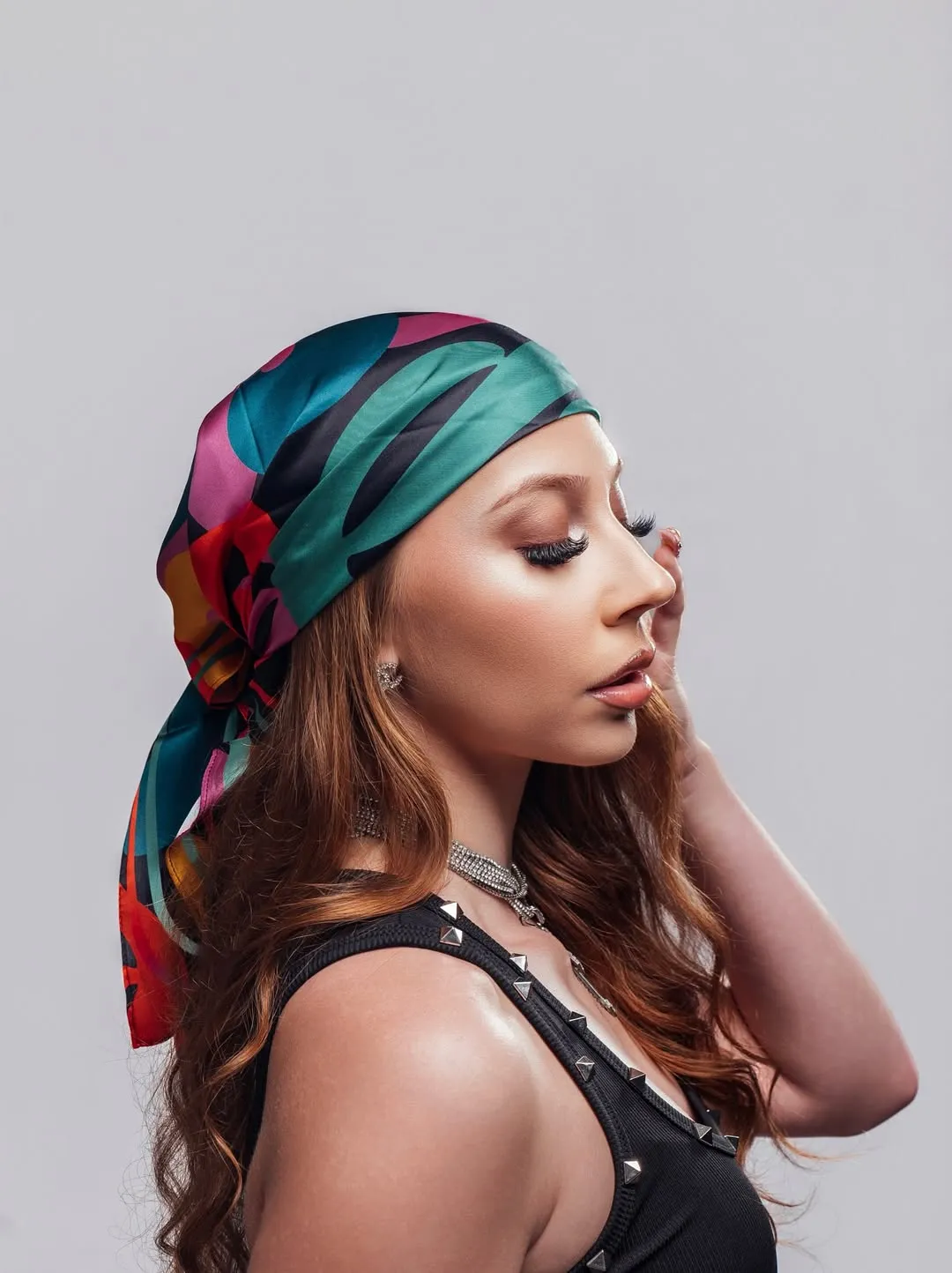 Woman with long brown hair in a colorful headscarf and black top poses with eyes closed against a gray background.